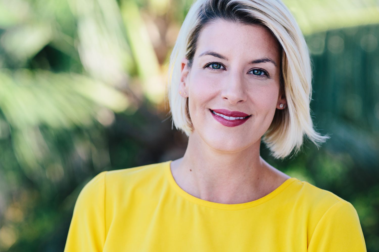 Professional headshot of a woman in a bright yellow top against a blurred outdoor background.