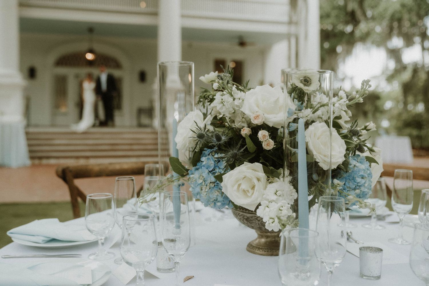 Elegant wedding centerpiece with white roses and light blue hydrangeas on a table setting at an outdoor reception.