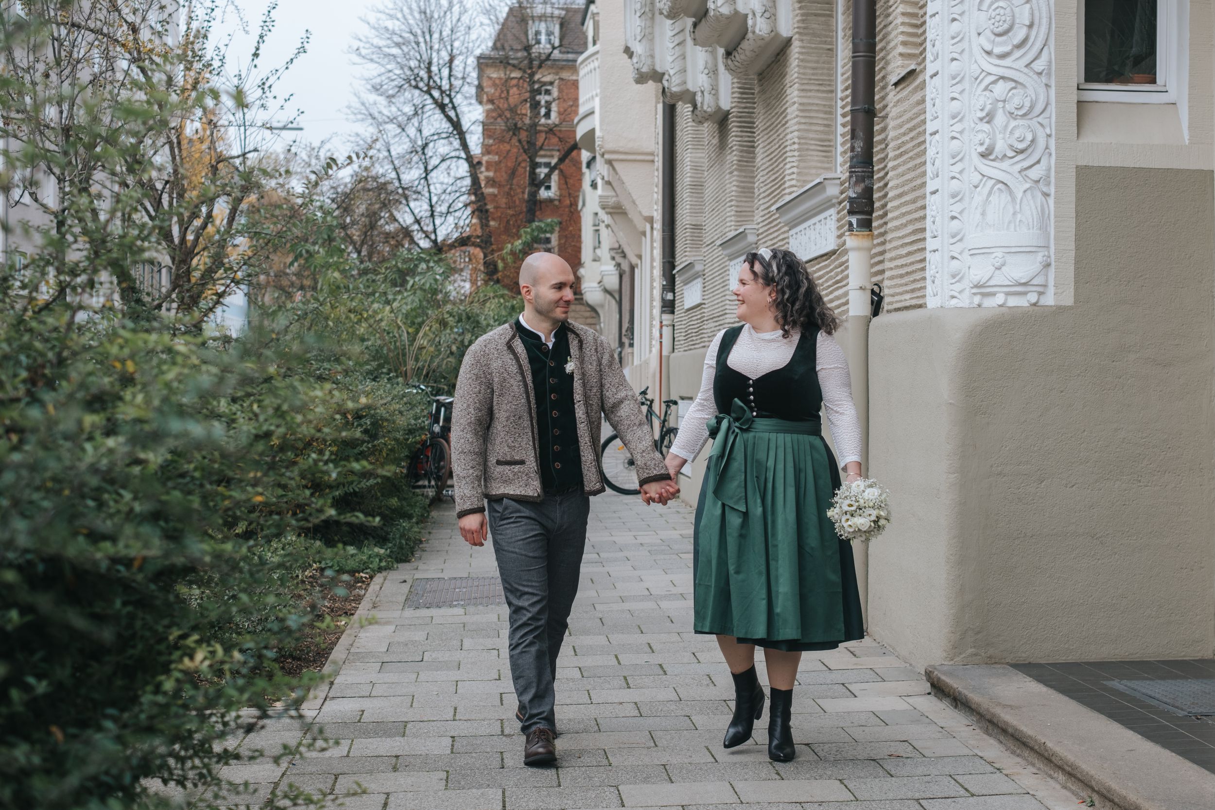 Hochzeitsfotografie München: Brautpaar geht Hand in Hand entlang eines Gehwegs vor einem Gebäude im städtischen Umfeld.