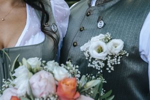 Hochzeitsfotografie: Nahaufnahme von traditioneller Hochzeitskleidung (Tracht) mit Blumenstrauß und Ansteckblume, fotografiert im Grünen.