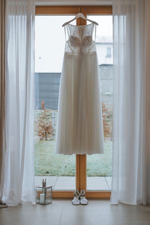 Hochzeitsfotografie: Brautkleid hängt am Fenster zwischen Vorhängen, ruhige Detailaufnahme vor der Hochzeit.