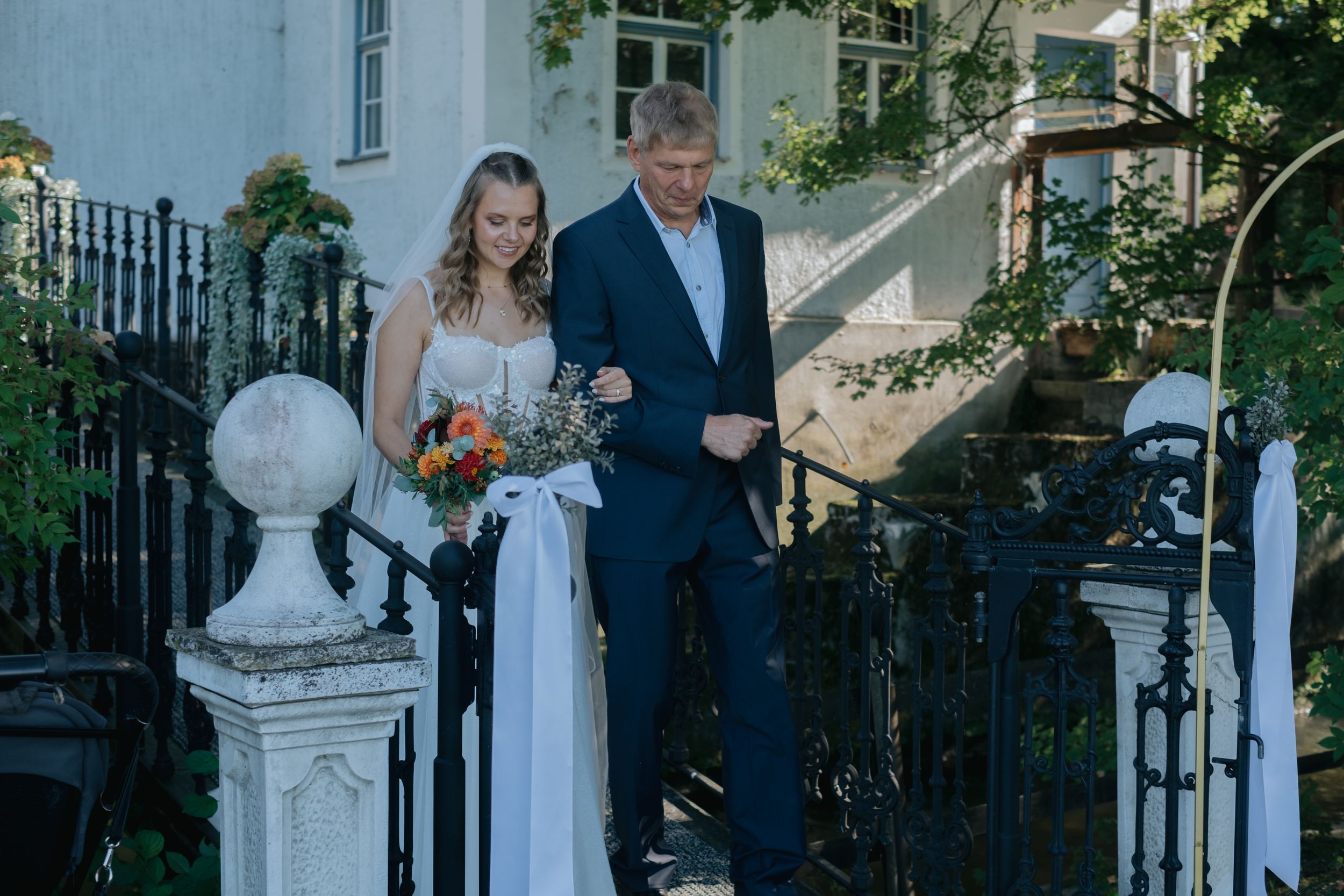 Hochzeitsfotografie Bayern: Braut geht mit Begleitung eine Treppe vor einem Gebäude hinunter, Beginn der Hochzeitsfeier.“