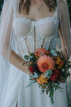 Hochzeitsfotografie: Detailaufnahme eines Brautkleids mit Spitzenoberteil und Schleier, die Braut hält einen farbenfrohen Blumenstrauß.