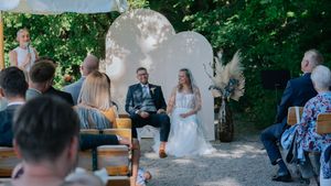 Hochzeitsfotografie: Freie Trauung im Grünen: Brautpaar sitzt vor einem dekorierten Traubogen, Gäste sitzen auf Holzbänken im Hintergrund.