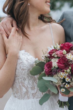 Hochzeitsfotografie: Detailaufnahme der Braut mit Spitzenkleid und Blumenstrauß, eine Hand liegt ruhig an der Schulter.