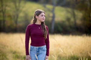 Young woman in burgundy sweater and jeans stands thoughtfully in golden autumn field at sunset.