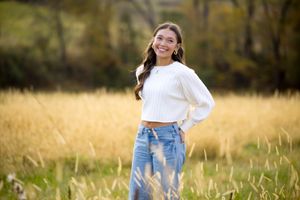 A woman in a white sweater and jeans walks through a golden autumn field at sunset with tall grass swaying in the breeze.