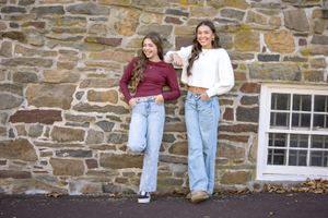 Two people sit casually on a rustic stone wall wearing jeans and contrasting burgundy and white tops.