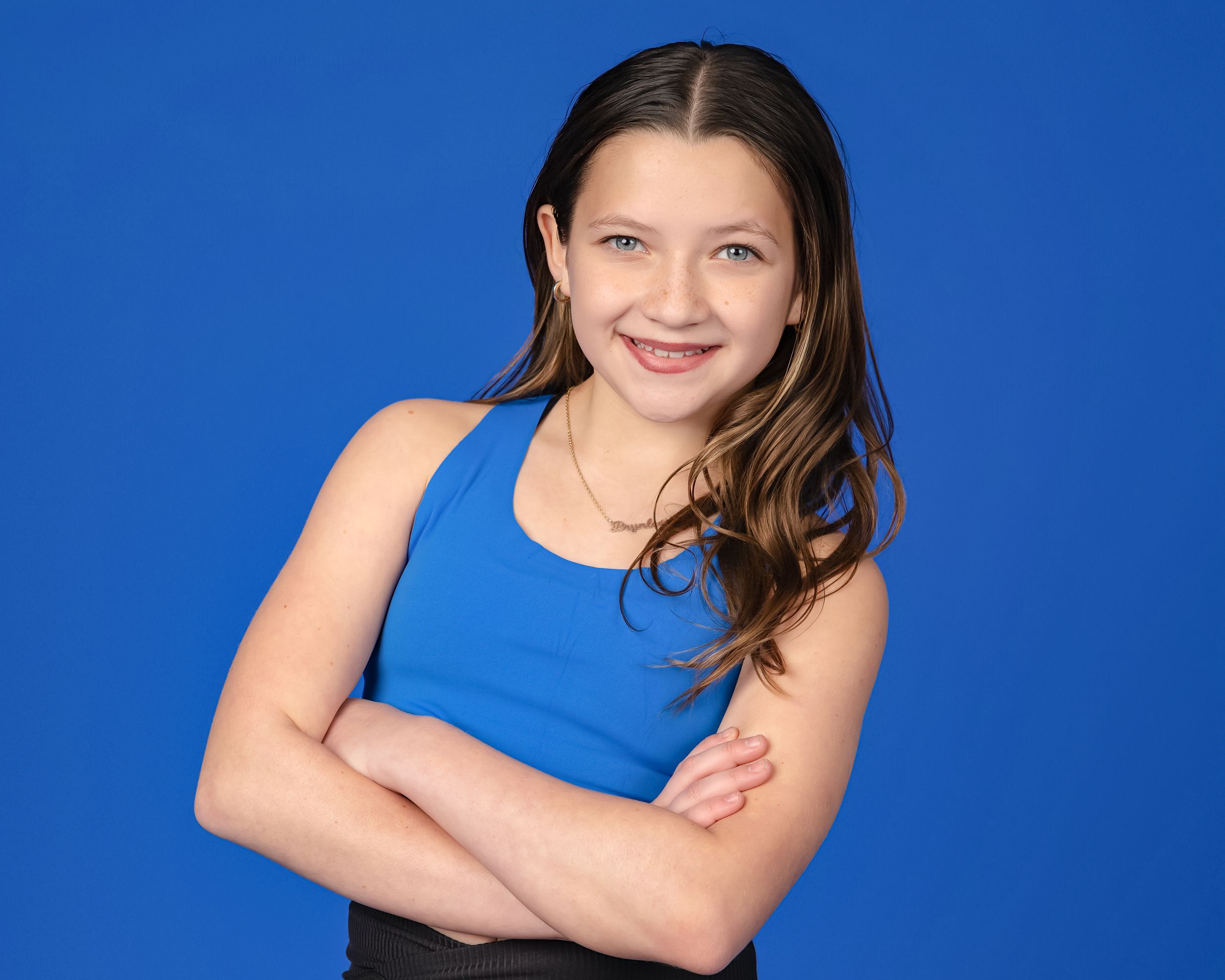 A person in a blue sleeveless top poses with arms crossed against a bright blue background.