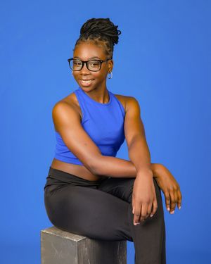 A person in a blue sleeveless top and black pants sits casually on a gray cube against a vibrant blue background.