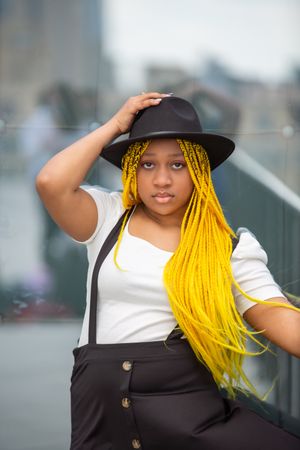 A person wearing a black hat and white top poses with bright yellow braids against a blurred outdoor background.