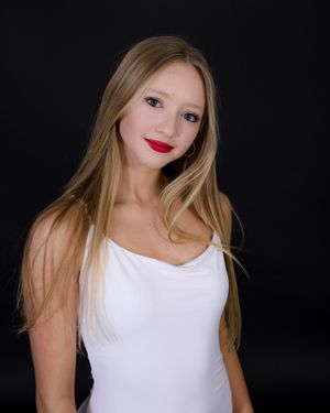 Portrait against dark background shows figure in white tank top with long blonde hair and bright red lipstick.