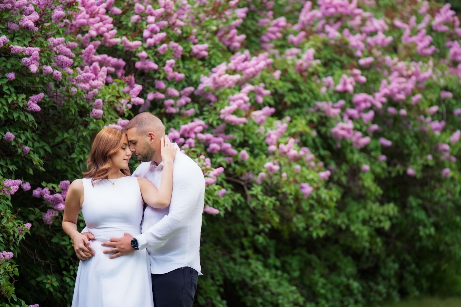 A romantic couple embraces in front of blooming pink lilac bushes in a spring garden setting.
