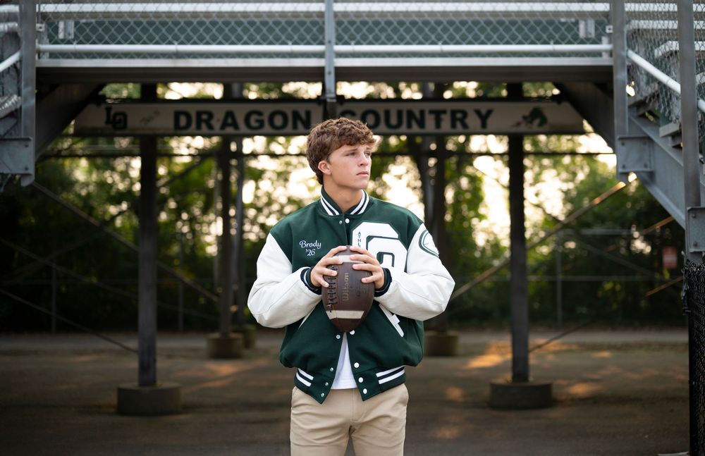 A person in a varsity jacket holds a football in front of metal bleachers at dusk.
