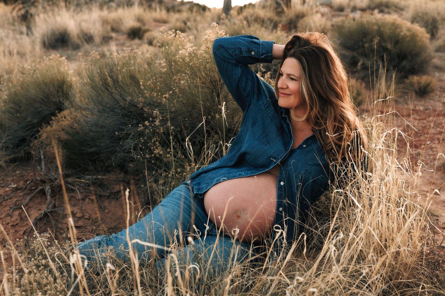 Maternity session, woman in white dress standing by a fence at golden hour with towering canyon rock formations behind her.