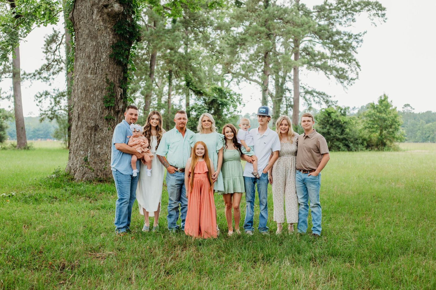 Family group portrait outdoors with coordinated outfits standing together in grassy field with trees.