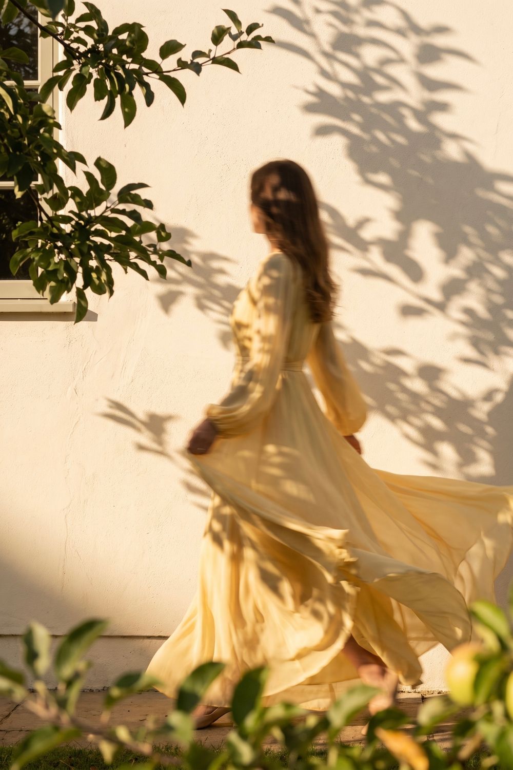 Woman in flowing yellow dress walking near white wall with leaf shadows in warm golden sunlight.