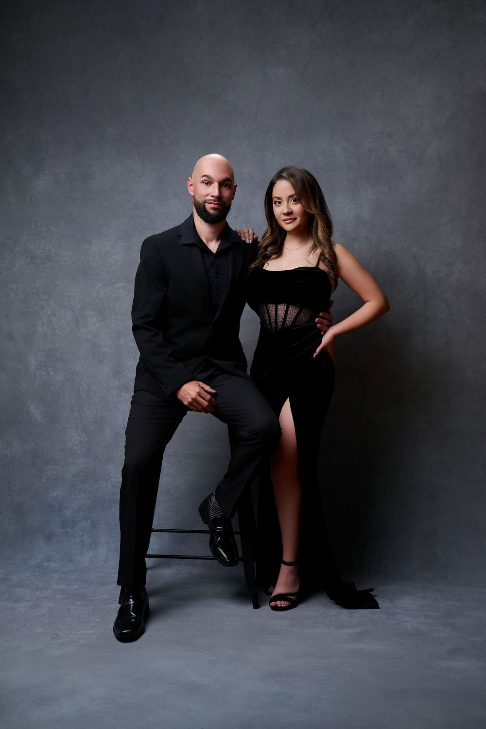 A couple in elegant black outfits poses against a gray backdrop, man in suit seated, woman in slit gown standing beside him.