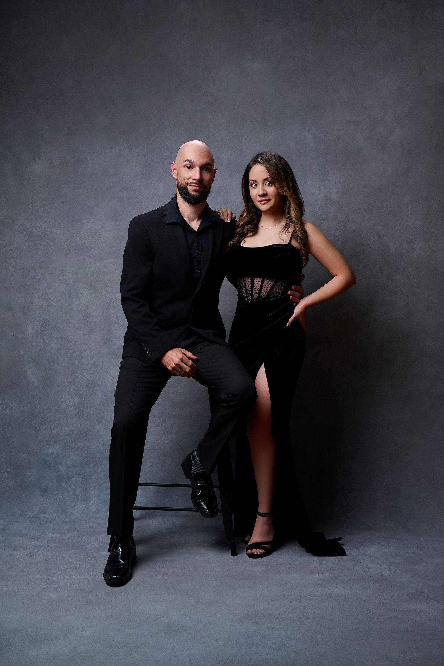 A couple in elegant black outfits poses against a gray backdrop, man in suit seated, woman in slit gown standing beside him.
