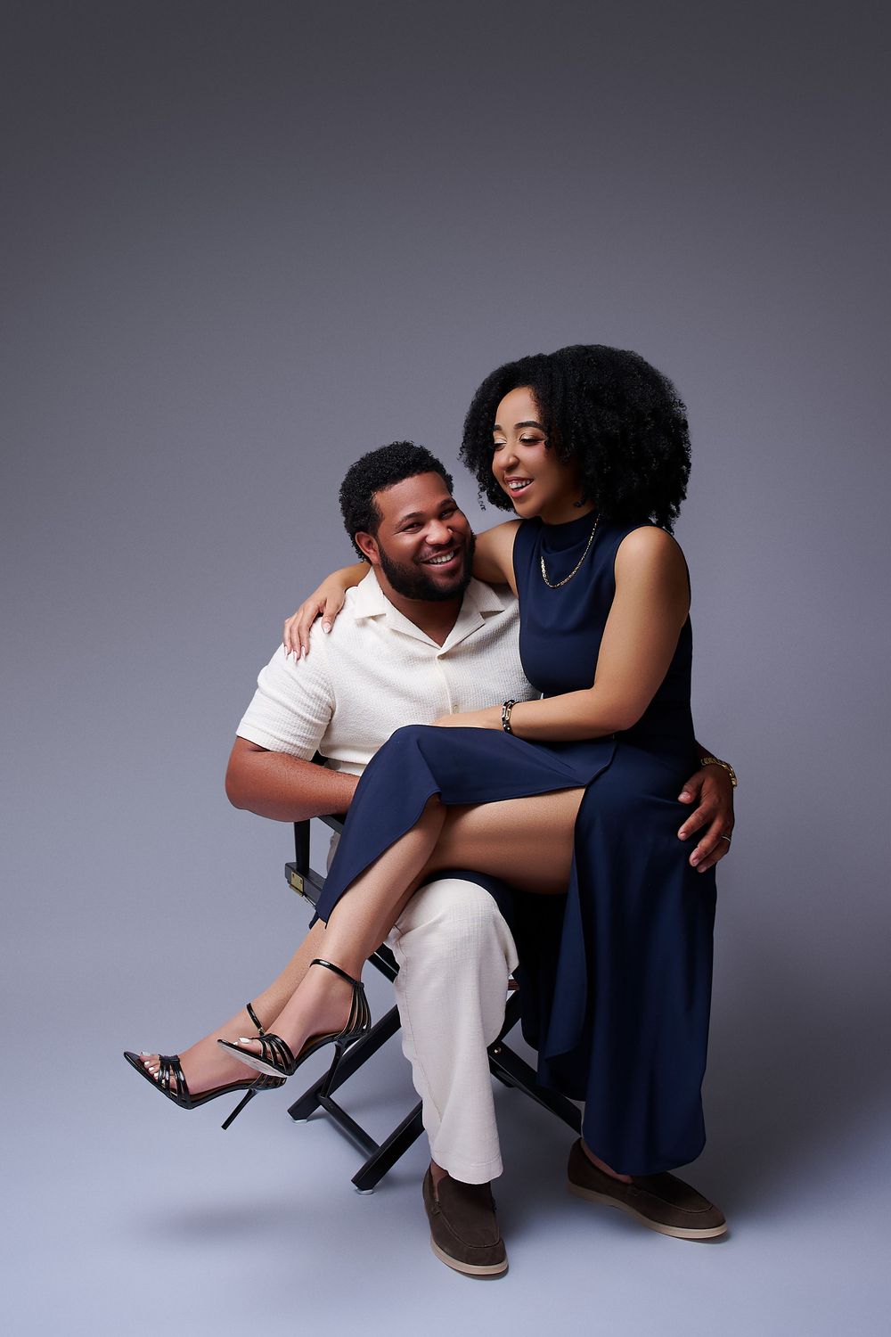 A smiling couple poses together, woman in navy dress sitting on man's lap in white outfit, studio portrait. NJ Studio engagement photos
