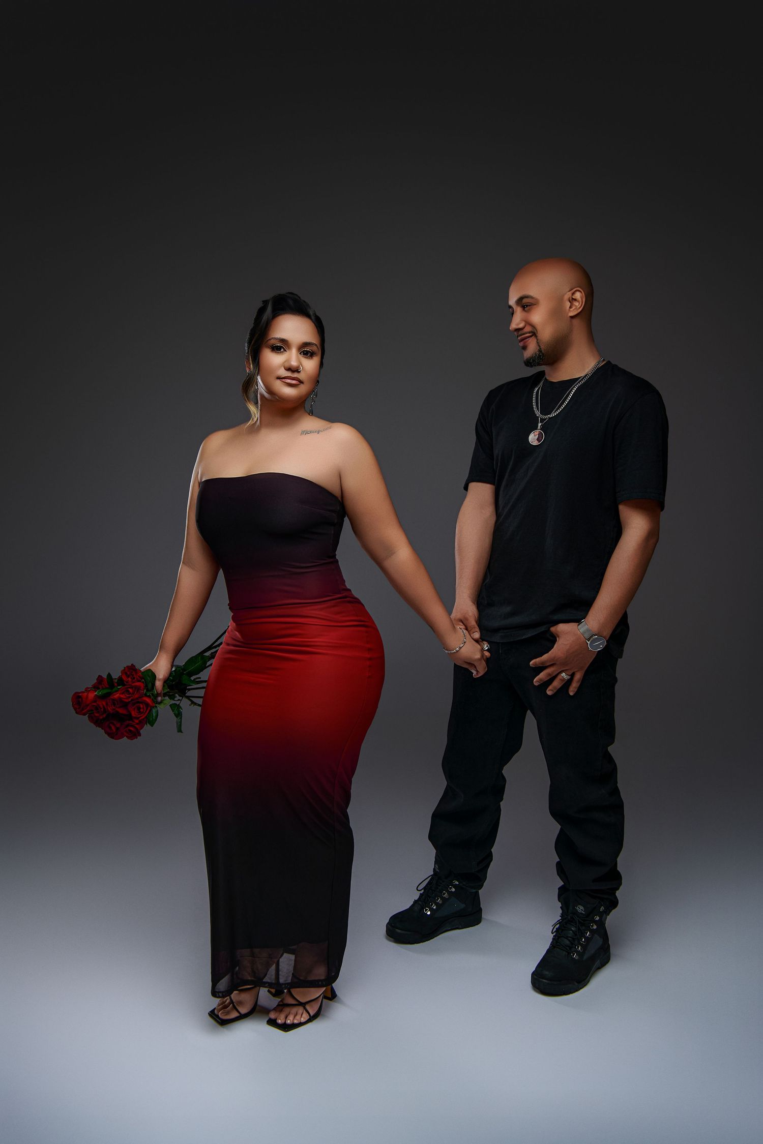 Couple holds hands in a studio setting, with woman in red ombre dress holding roses and man wearing all black.