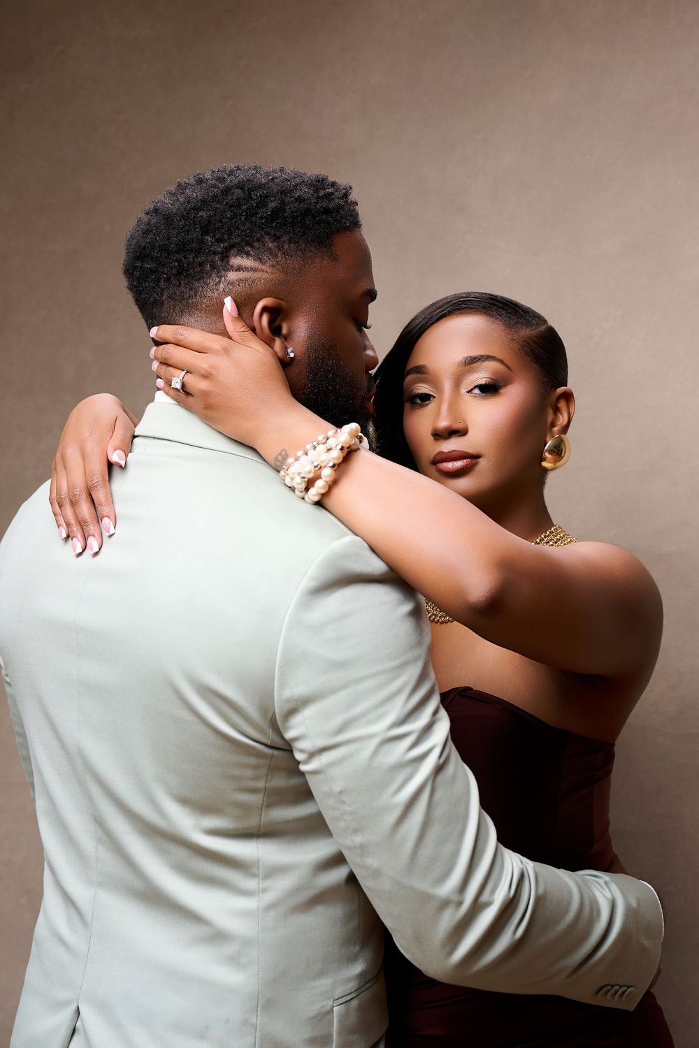 Elegant couple embracing in studio, woman in strapless brown dress with gold jewelry, man in light suit.