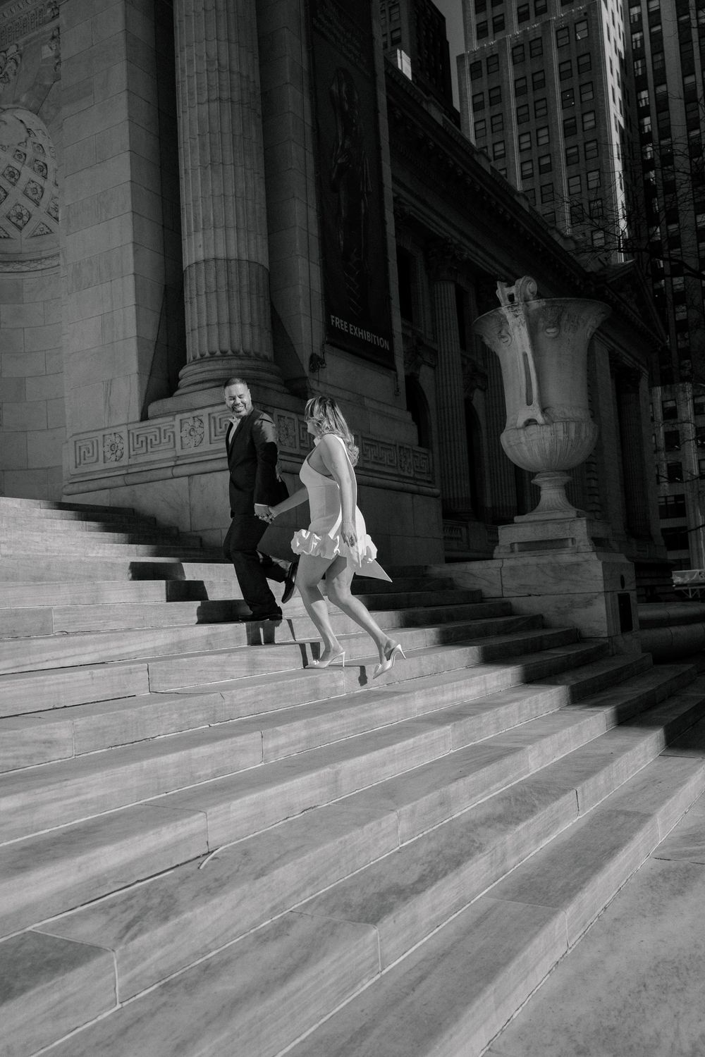 Black and white photo of a couple in wedding attire running up grand stone steps of a classic building.