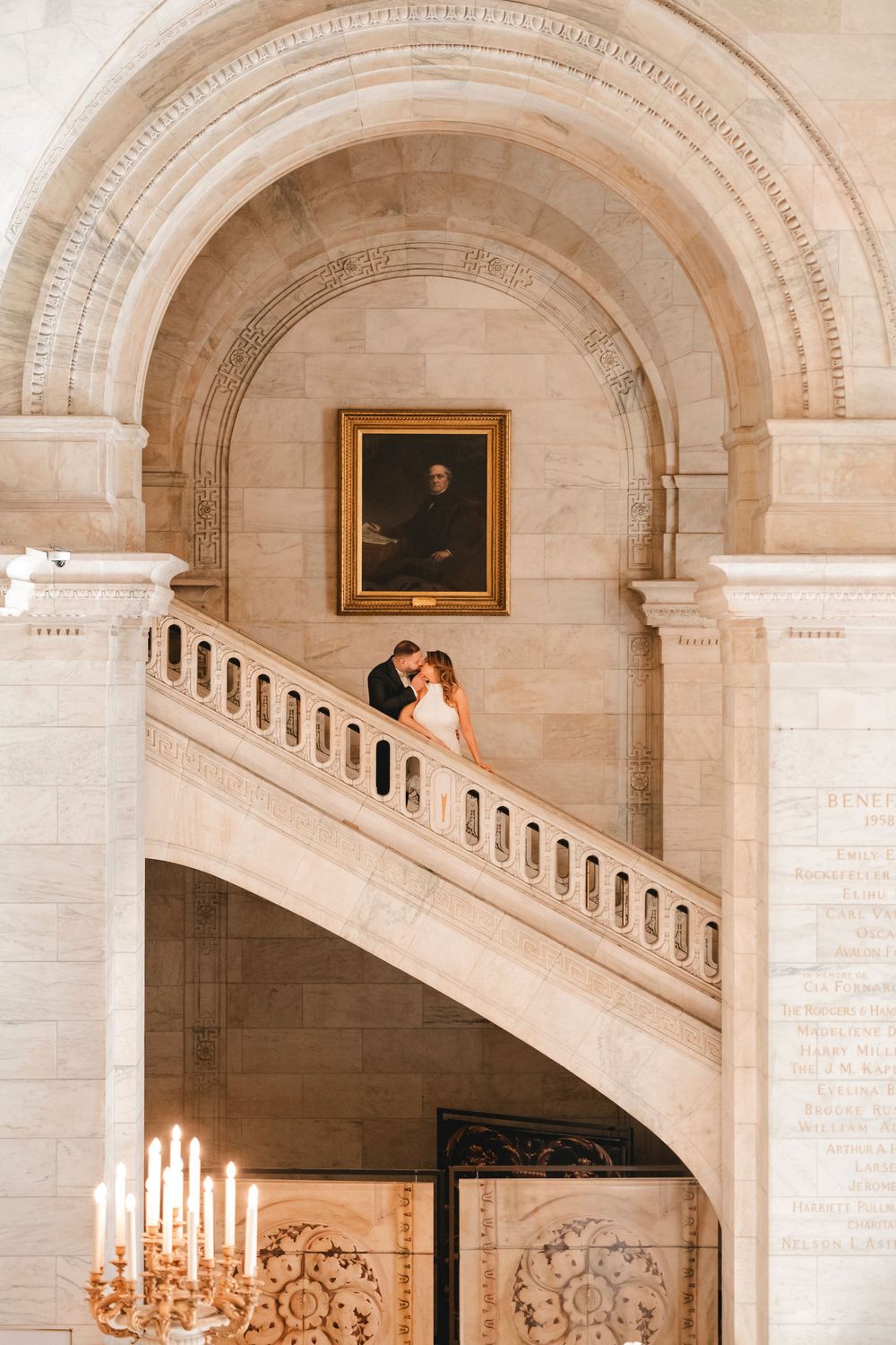 Bride and groom embrace on grand marble staircase beneath arched ceiling and portrait in elegant historic venue.