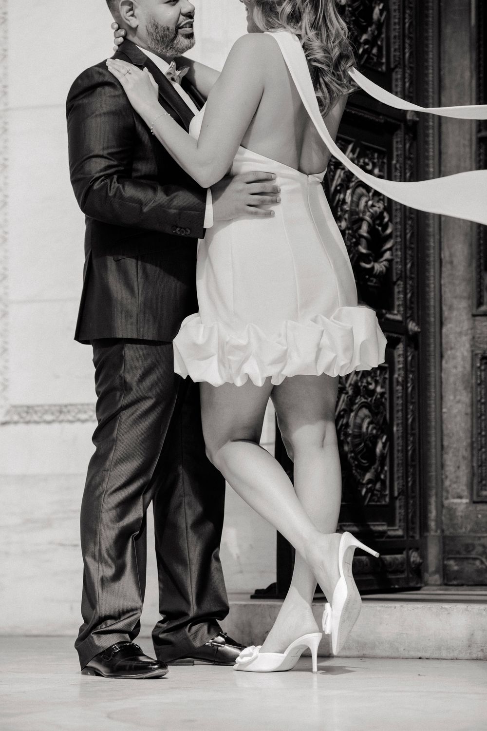 Black and white photo of a couple embracing, bride in short white dress and heels, groom in dark suit.