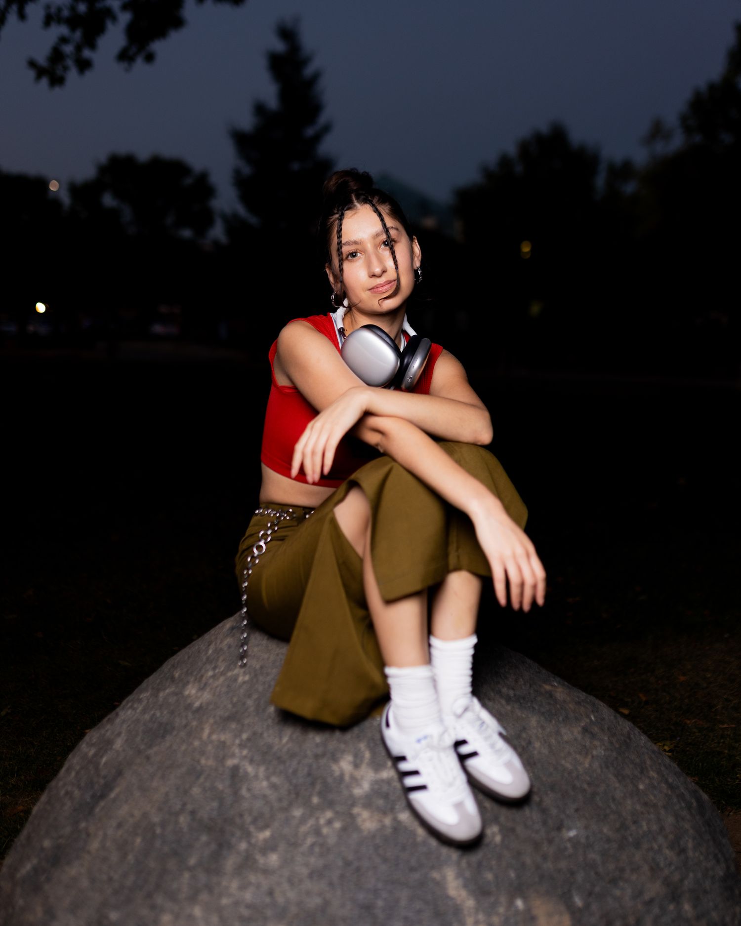 Person in red crop top and olive pants sits on large rock at dusk wearing white sneakers and headphones around neck.