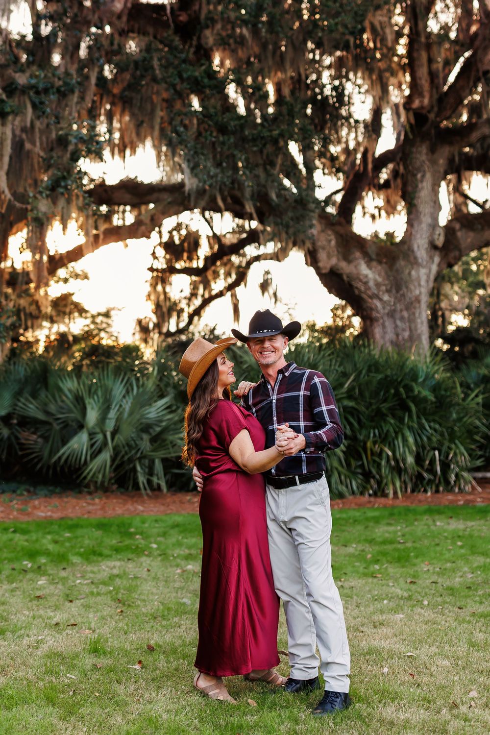 A romantic couple shares an intimate moment under Spanish moss draped oak trees at sunset in a park setting.