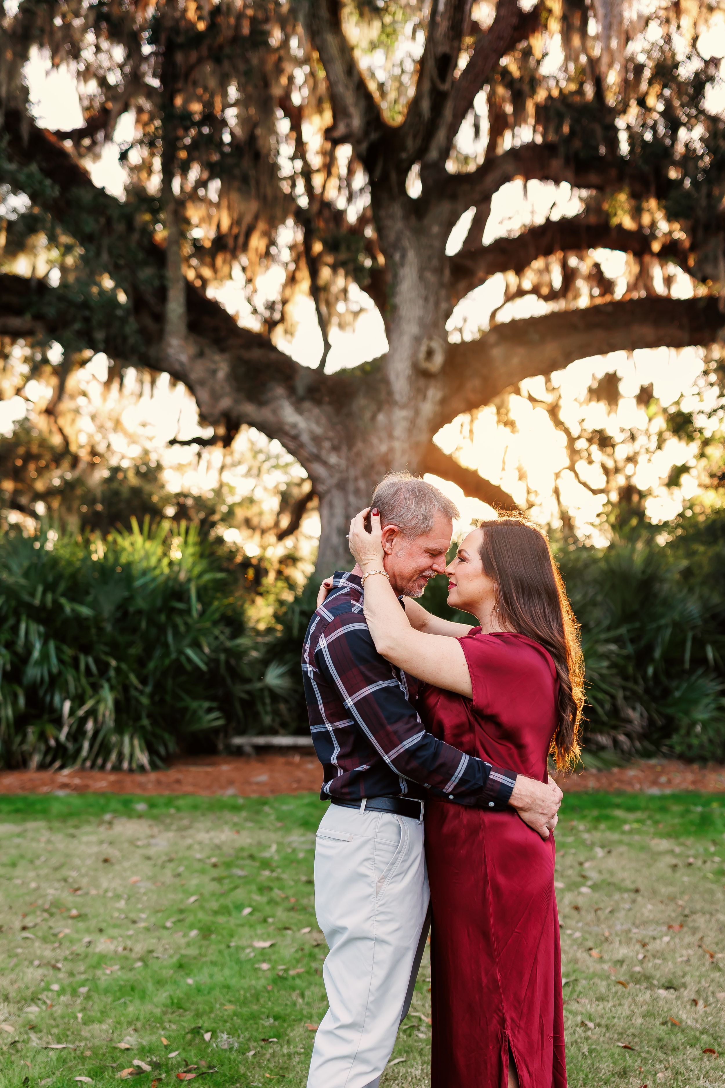 A couple in red and plaid embraces beneath a large oak tree during sunset in a park setting.