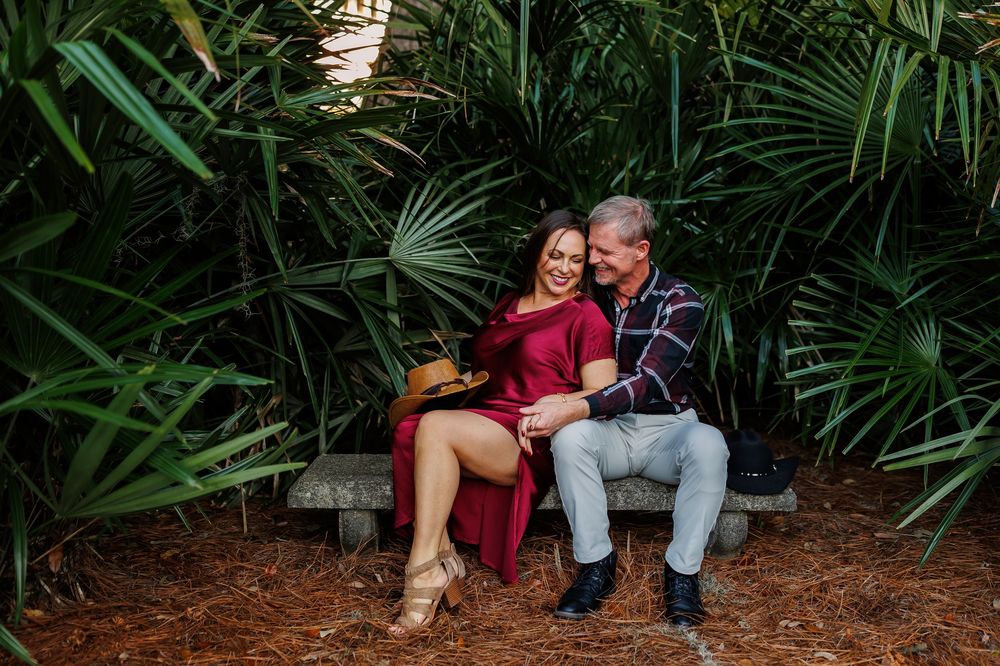 Couple sitting together on wooden bench surrounded by lush tropical plants and greenery in garden setting.