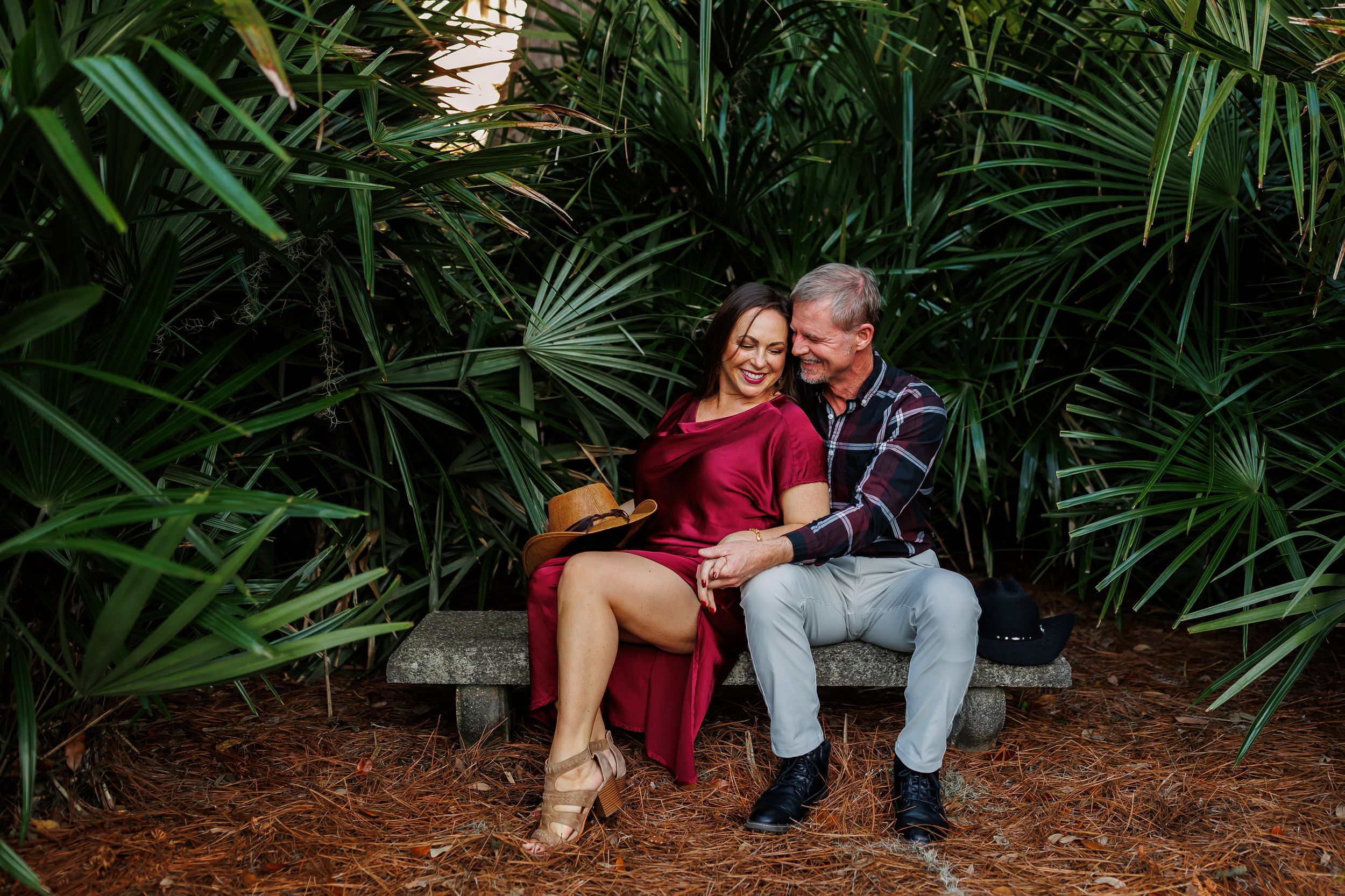 Couple sitting together on wooden bench surrounded by lush tropical plants and greenery in garden setting.