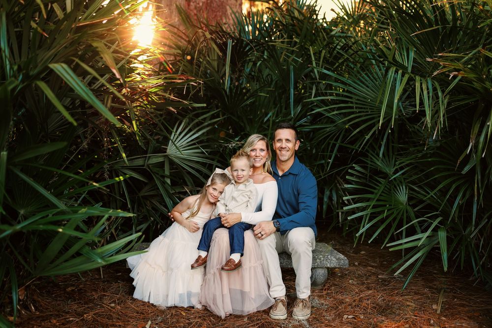 A family sits together on a bench surrounded by lush palm fronds at sunset in a tropical garden setting.