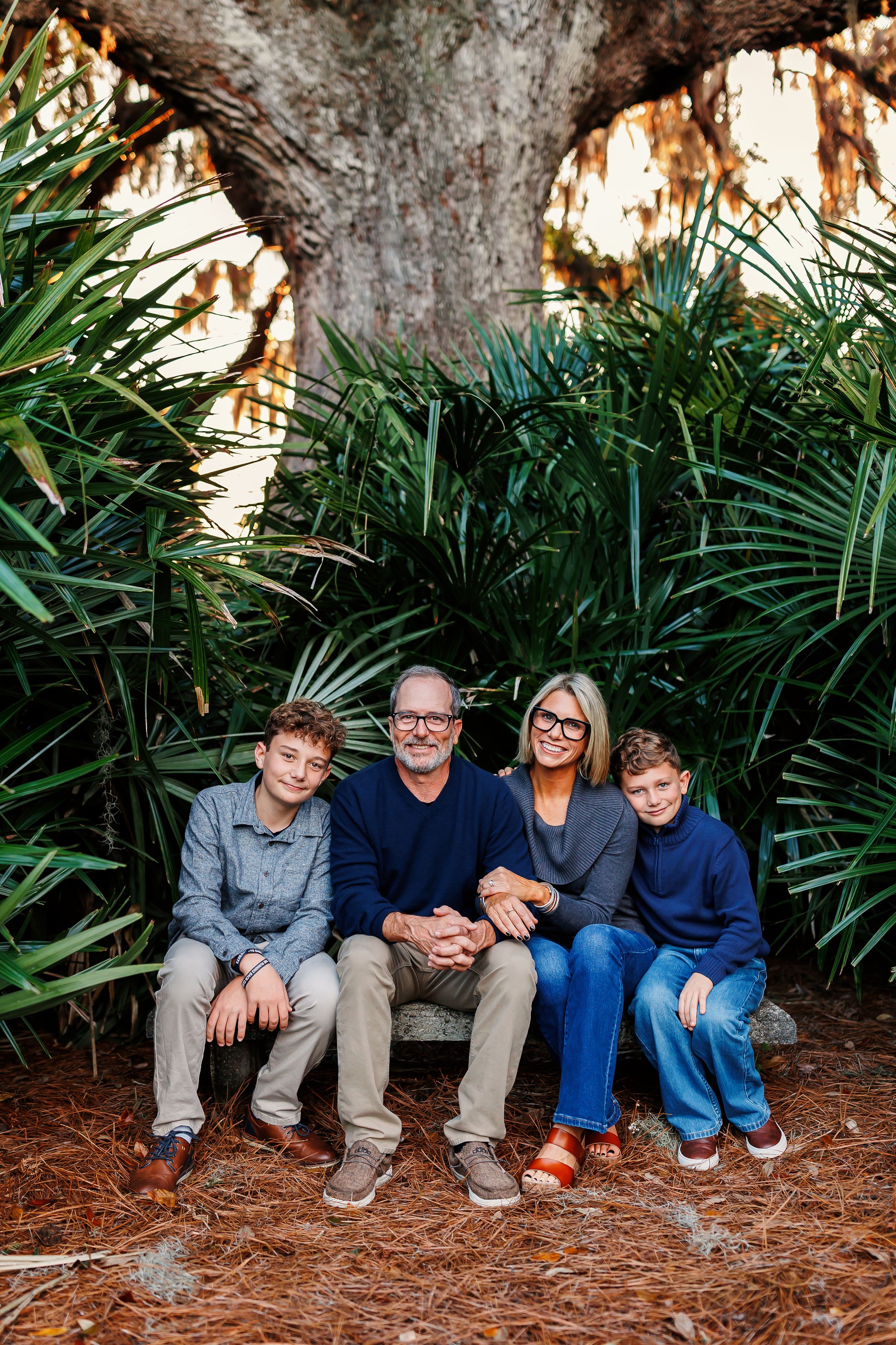 A group sits together on a wooden bench surrounded by lush tropical palm fronds and greenery in an outdoor setting.