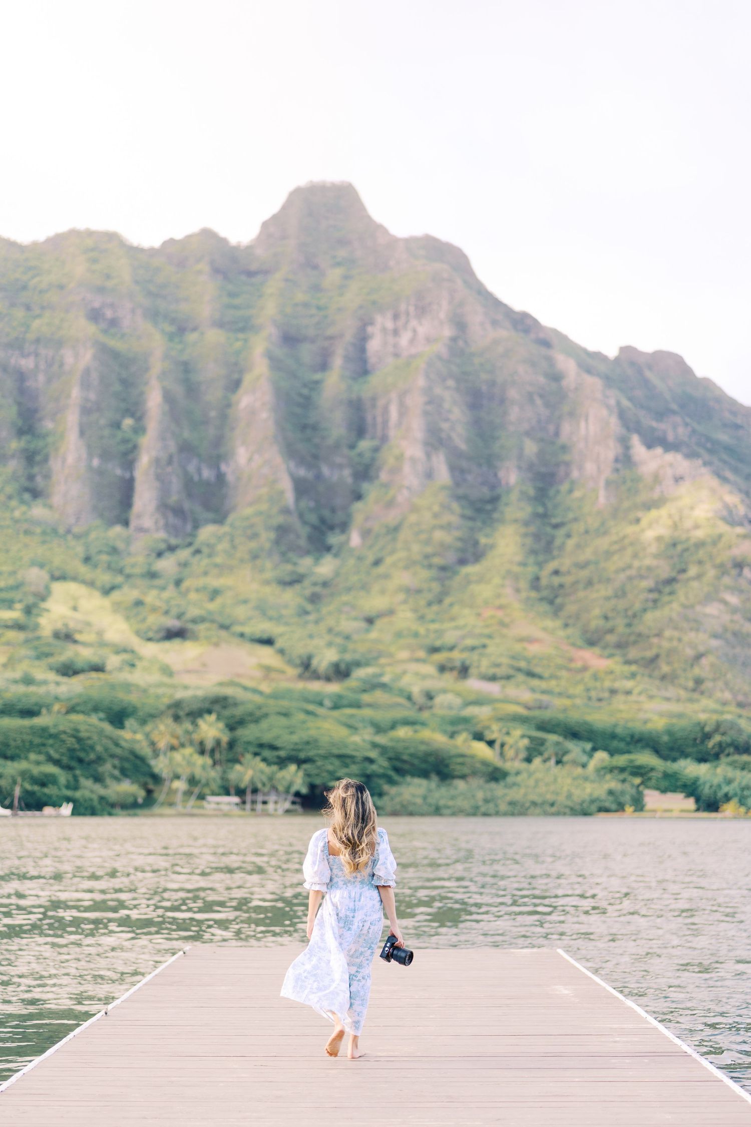 A person in white dress walks along wooden pier with dramatic Hawaiian mountain landscape in background.