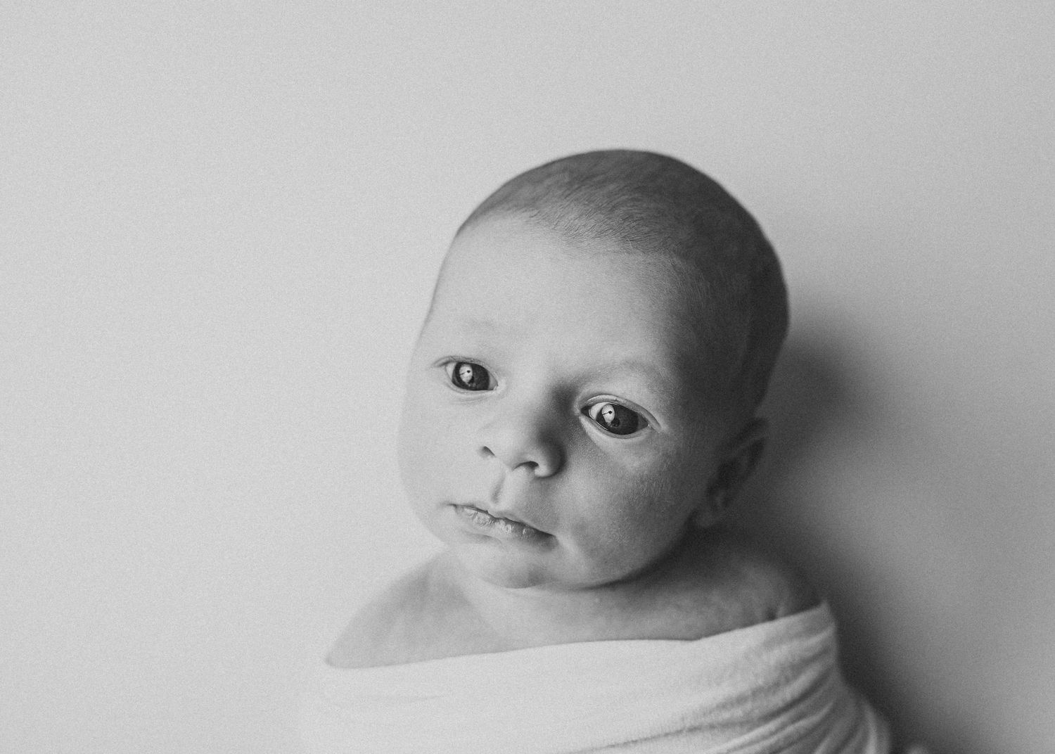 Black and white newborn portrait of swaddled baby with alert eyes on white background.