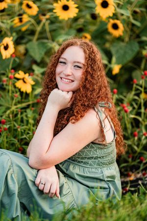 Beautiful portrait of long curly red hair in a sage green dress sitting among bright blooming sunflowers in a garden.
