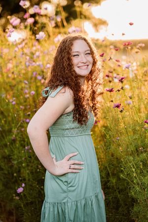 A curly-haired person in a mint green dress smiles in a golden wildflower field at sunset.