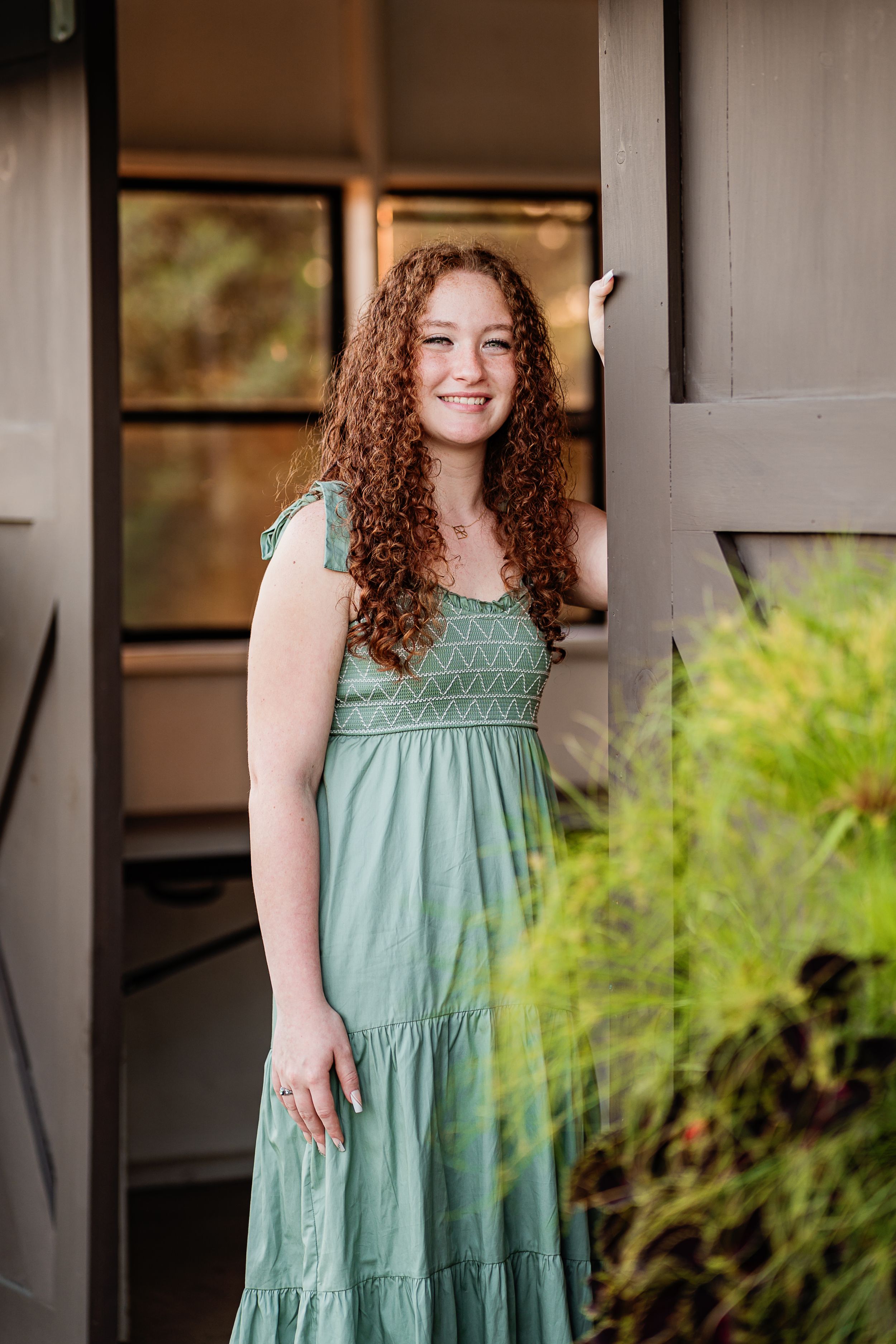 A smiling person in a sage green tiered maxi dress stands in a doorway with natural greenery visible outside.