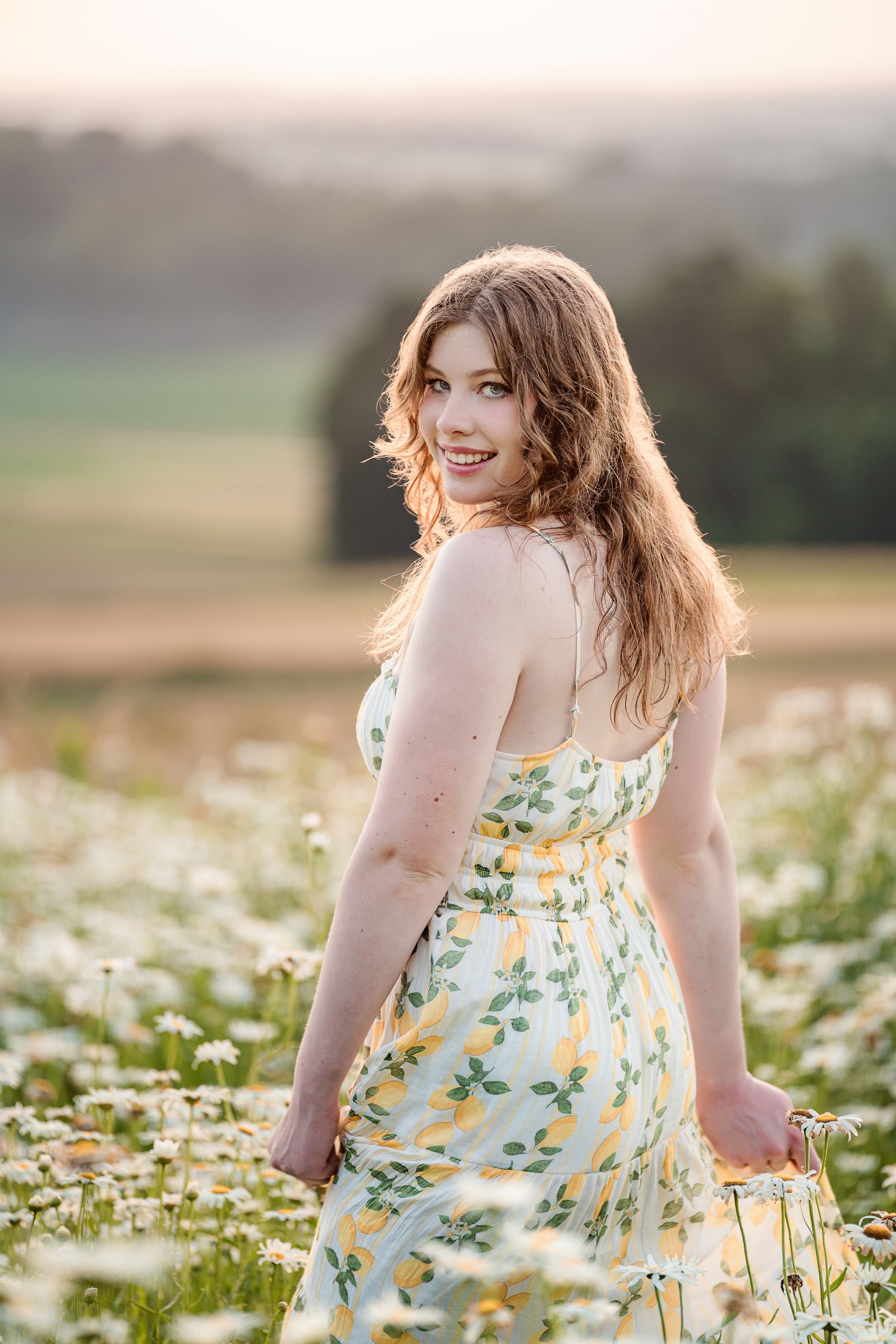 Person in a floral yellow sundress poses in a field of daisies during golden hour sunset.