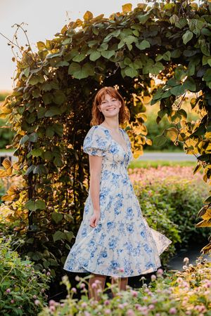 A garden scene at sunset with someone in a blue and white floral dress standing under a leafy arch.