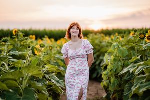 A person wearing a floral dress stands in a field of tall sunflowers during sunset.