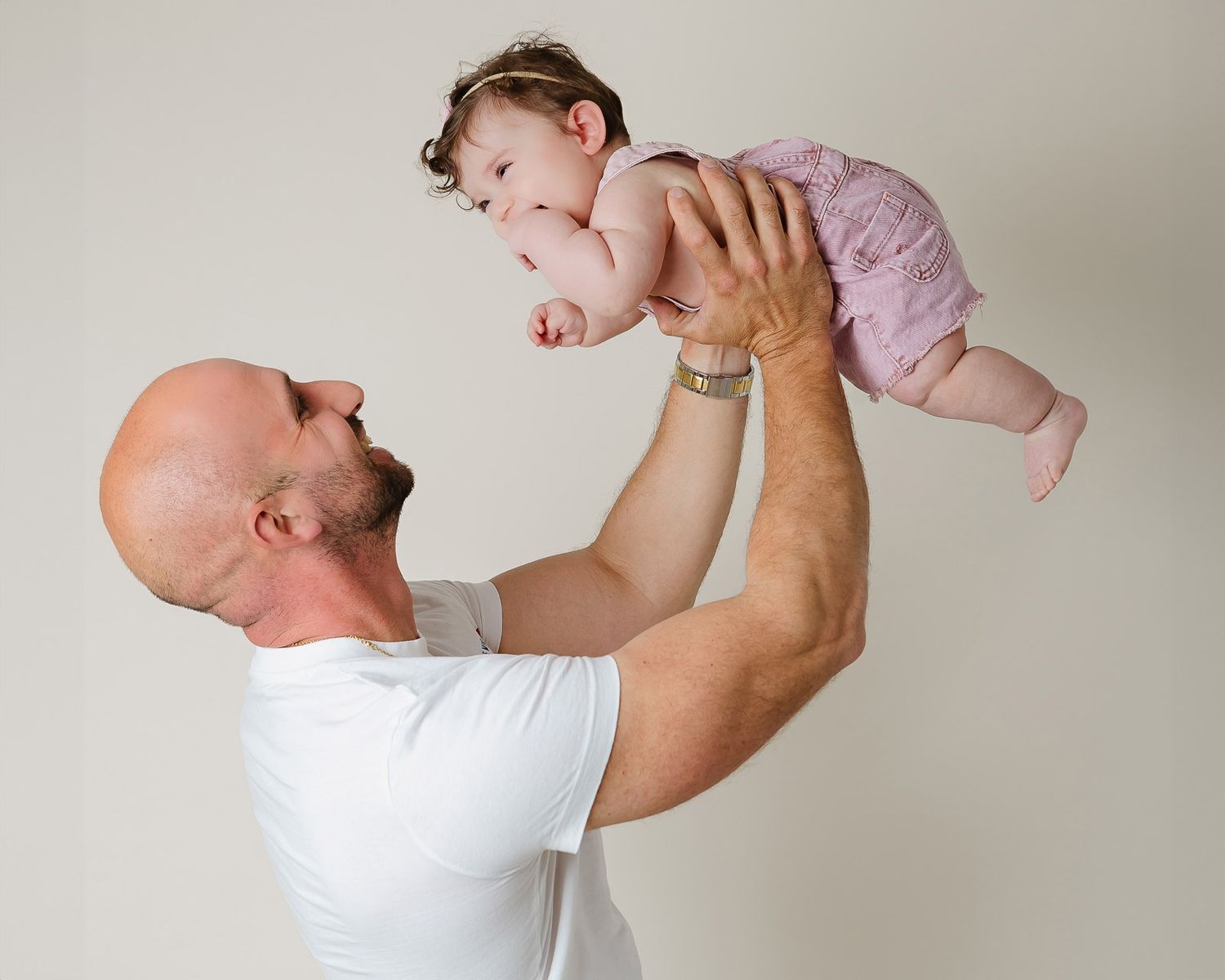 A person in a white t-shirt lifts a baby wearing pink clothing against a light colored background.