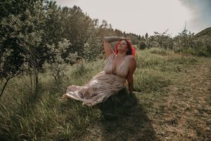 A woman with red hair in a flowing beige dress sits gracefully on a grassy hillside surrounded by trees.