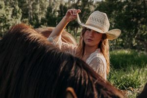 Woman in white lace dress and straw cowboy hat posing with a dark horse in a green outdoor setting.