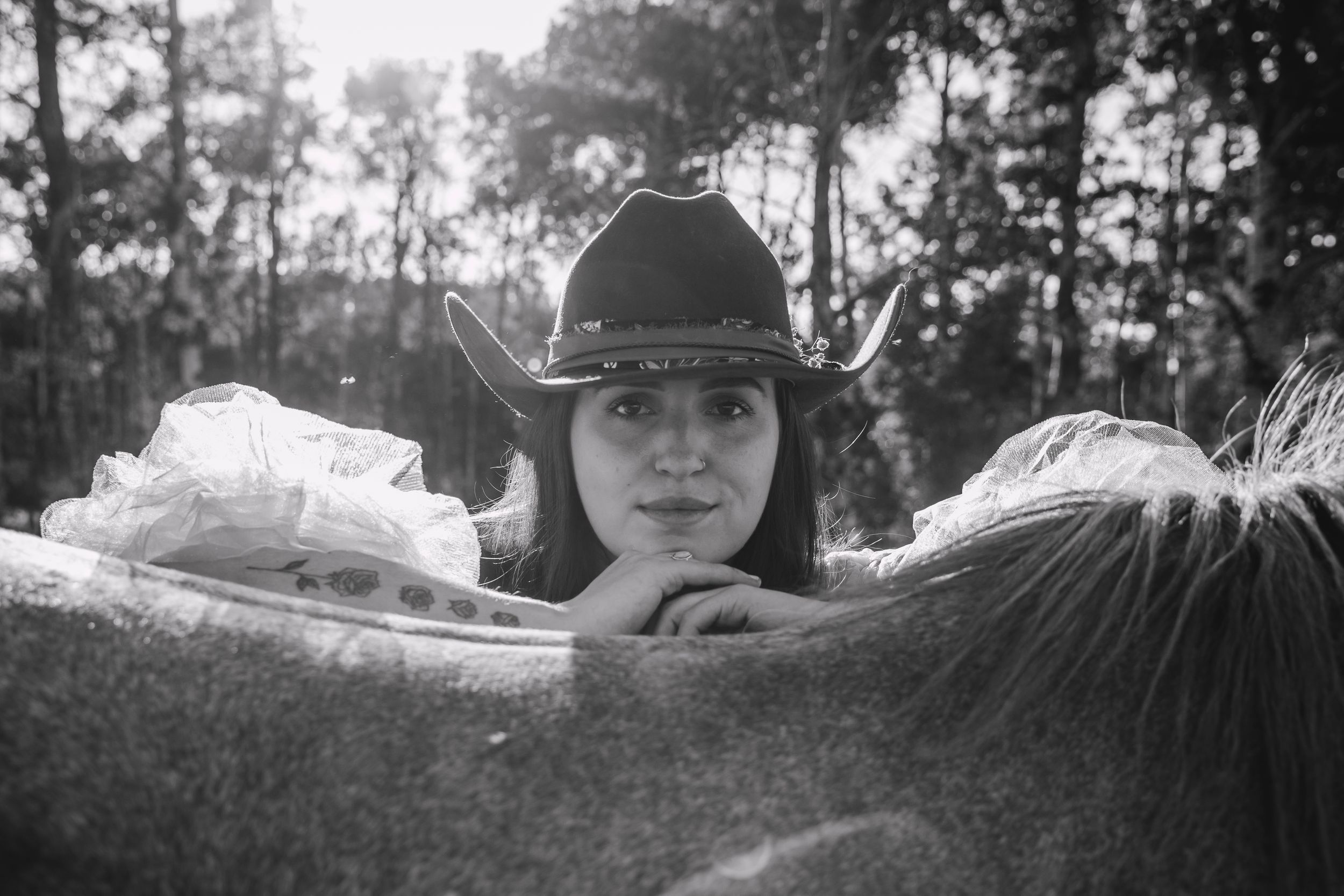 Black and white photo of a woman in a cowboy hat resting her arms on a horse, with trees in the background.