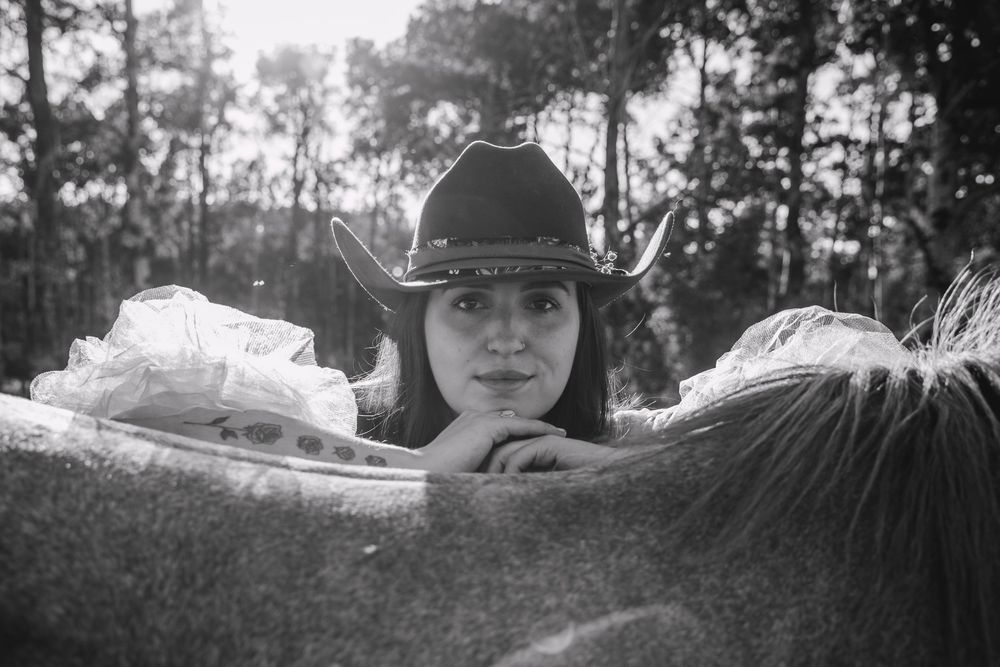 Black and white photo of a woman in a cowboy hat resting her arms on a horse, with trees in the background.