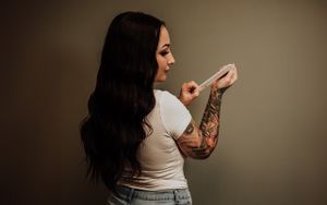 A woman with long dark hair and tattoos stands in profile against a neutral background wearing a white shirt.
